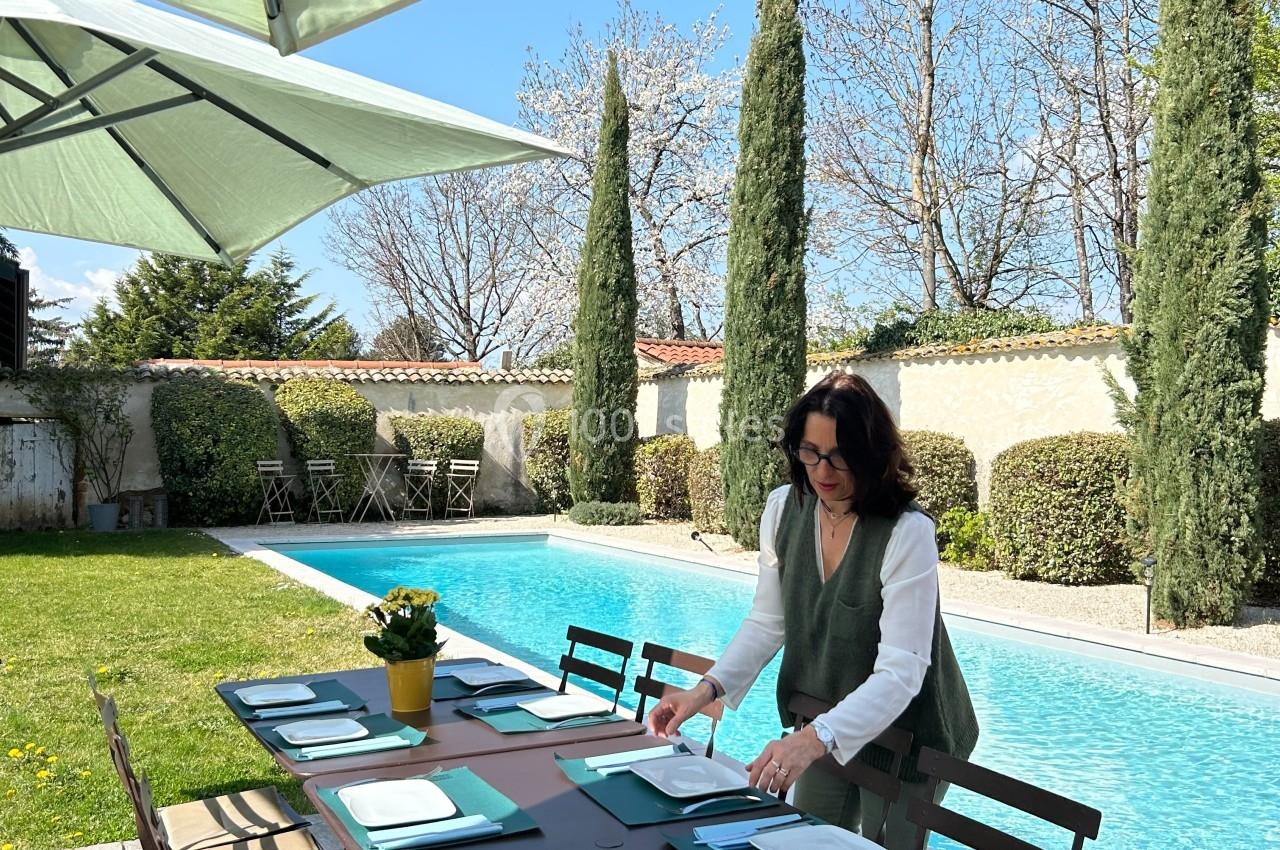 Une femme prépare une table extérieure dressée près d'une piscine, entourée de parasols et de végétation.
