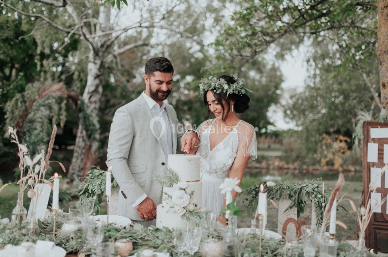 Un couple coupe un gâteau de mariage dans un décor extérieur avec une table décorée de fleurs et de bougies.