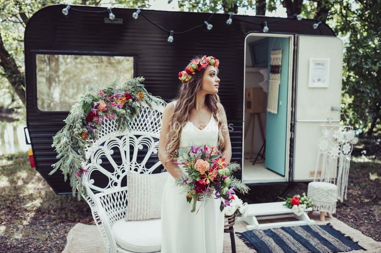 Femme en robe blanche tenant un bouquet coloré devant une caravane décorée de fleurs dans un cadre naturel.