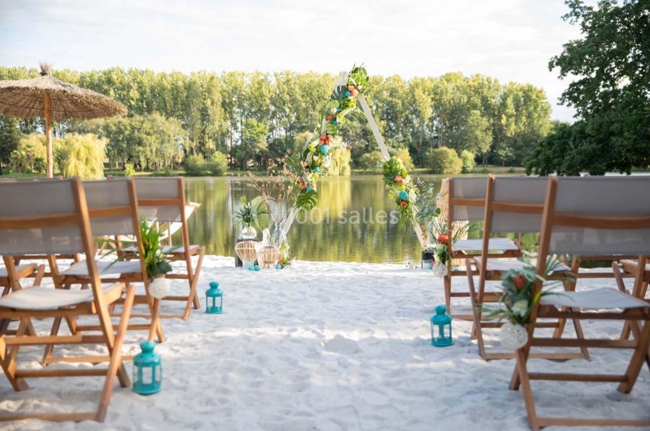 Chaises en bois disposées sur du sable face à une arche décorée, avec un lac et des arbres en arrière-plan.