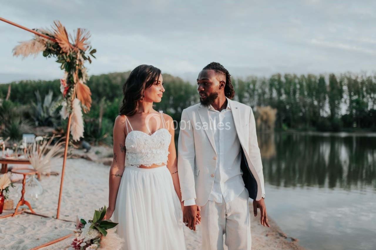 Un couple en tenue de mariage marche main dans la main sur une plage près d'un lac, décoré pour une cérémonie.