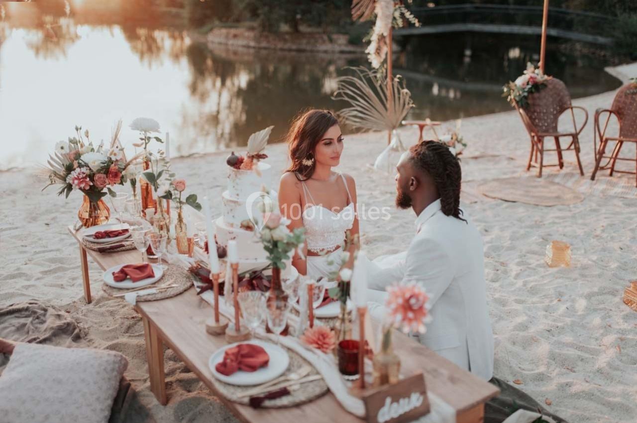 Un couple assis à une table décorée sur une plage au coucher du soleil, entouré de fleurs et de bougies.