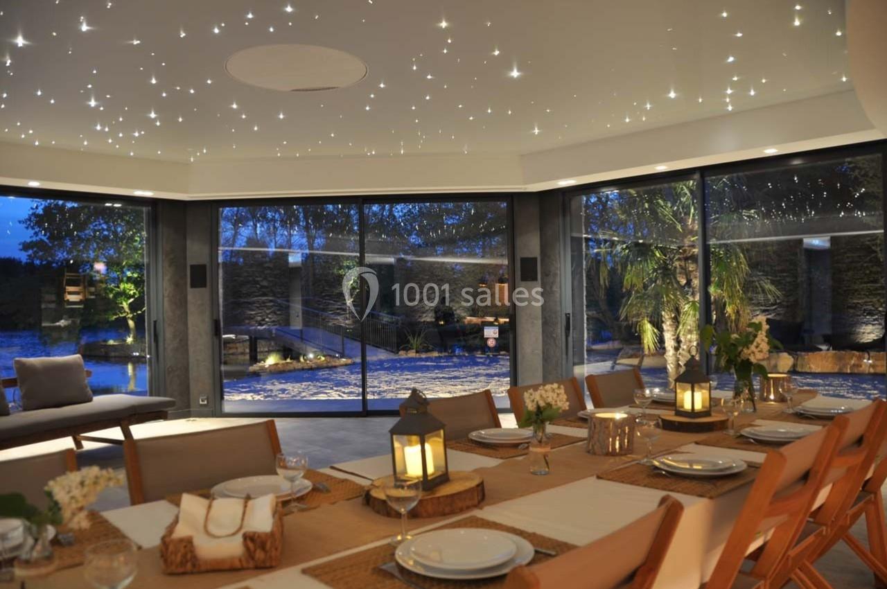 Salle à manger élégante avec grande table dressée, éclairage tamisé et vue sur un jardin nocturne à travers des baies…