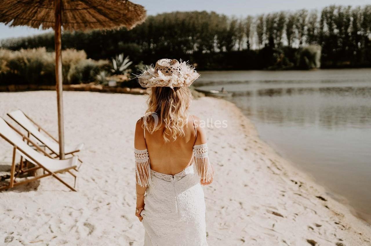 Une femme en robe de mariée dos nu marche sur une plage de sable près d'un lac, sous un parasol en paille.