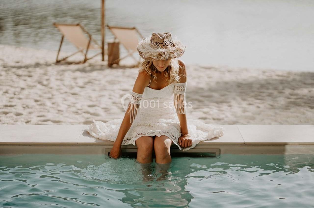 Une femme en robe blanche et chapeau assise au bord d'une piscine, les pieds dans l'eau, avec une plage en arrière-plan.