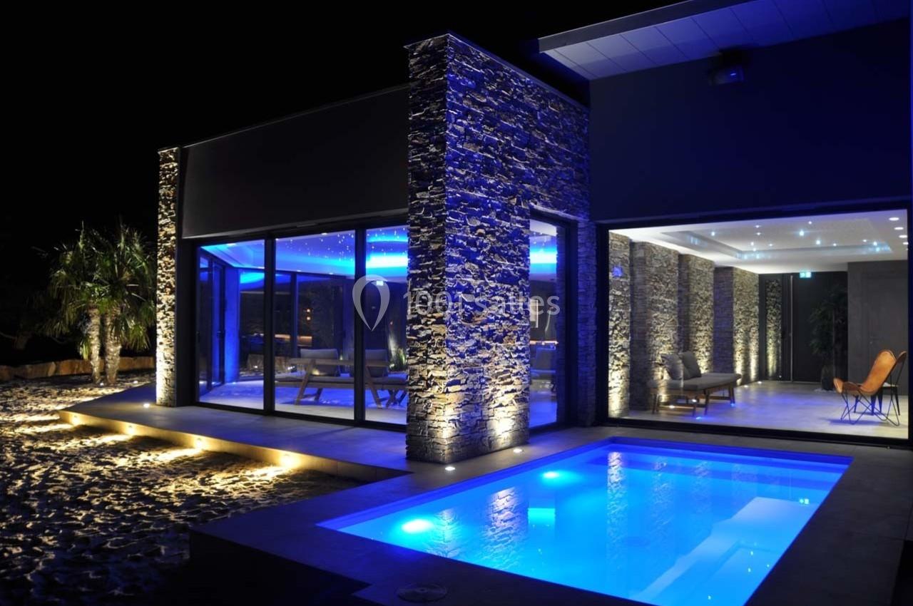 Maison moderne avec façade en pierre, éclairage bleu, grandes baies vitrées et piscine illuminée de nuit.