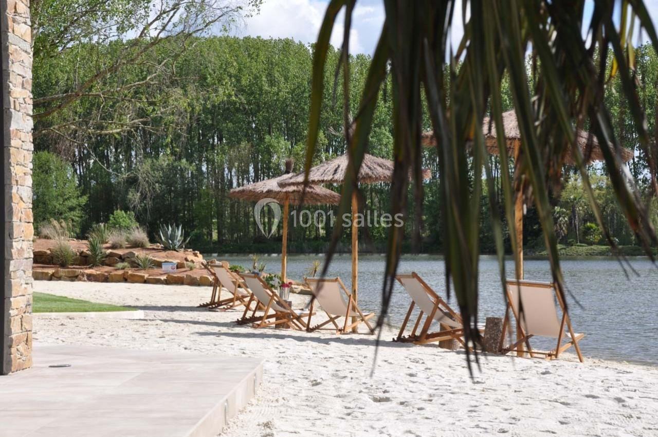 Chaises longues en bois disposées sur une plage de sable près d'un lac, entourée de végétation et d'arbres.