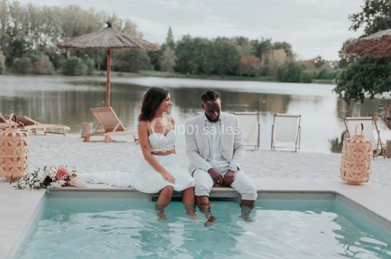 Un couple assis au bord d'une piscine, pieds dans l'eau, avec un lac et des transats en arrière-plan.