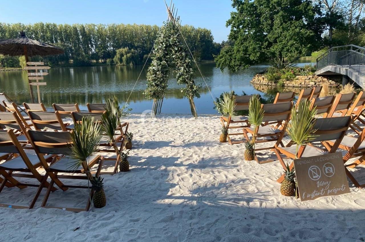 Chaises en bois disposées sur du sable face à une arche décorée, avec un lac et des arbres en arrière-plan.