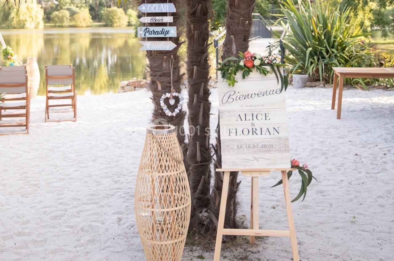 Panneau de bienvenue pour un mariage, posé sur un chevalet près de palmiers, avec des chaises et un lac en arrière-plan.
