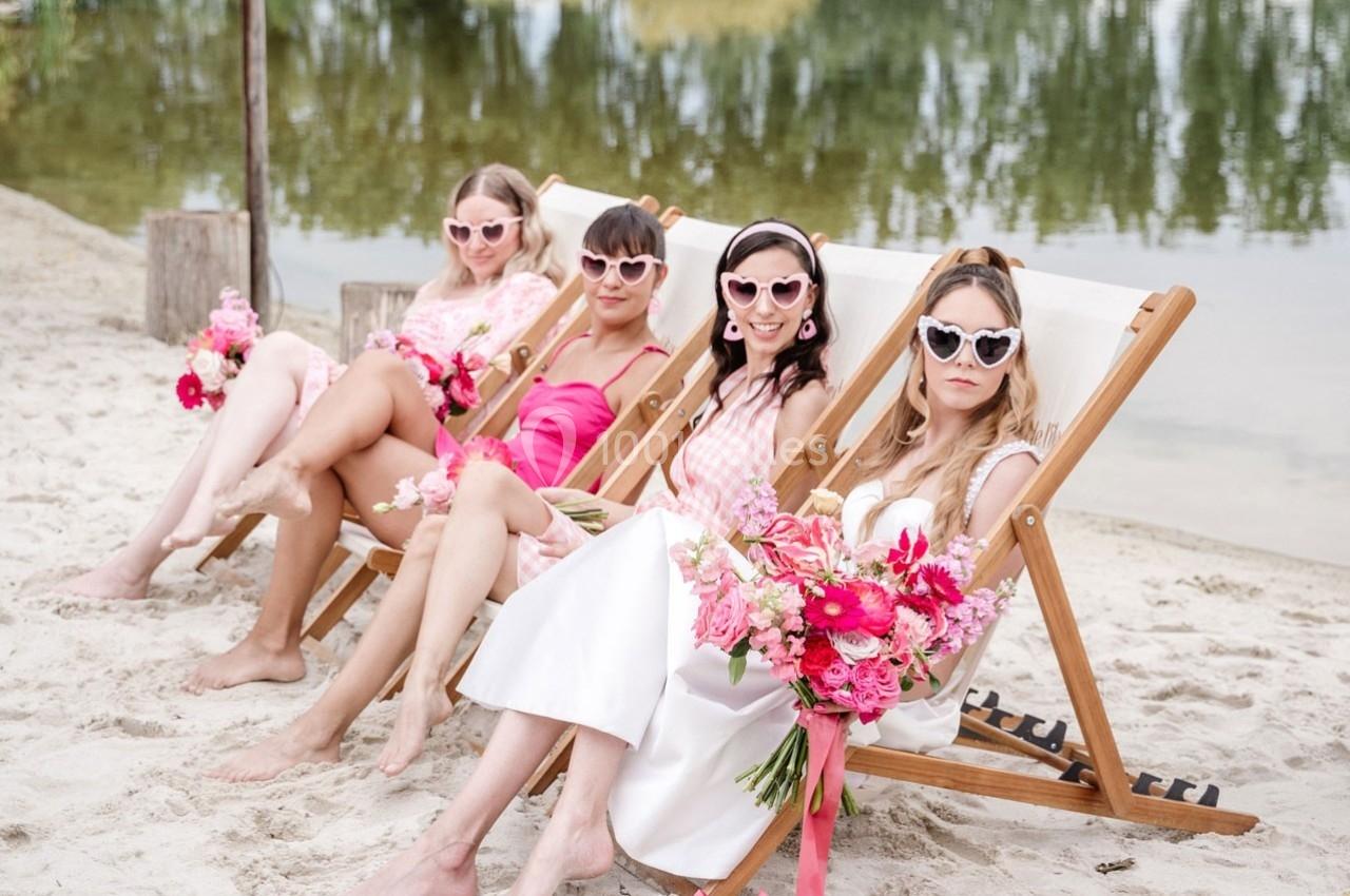 Quatre femmes assises sur des transats au bord d'une plage, portant des lunettes de soleil et tenant des bouquets de fleurs…