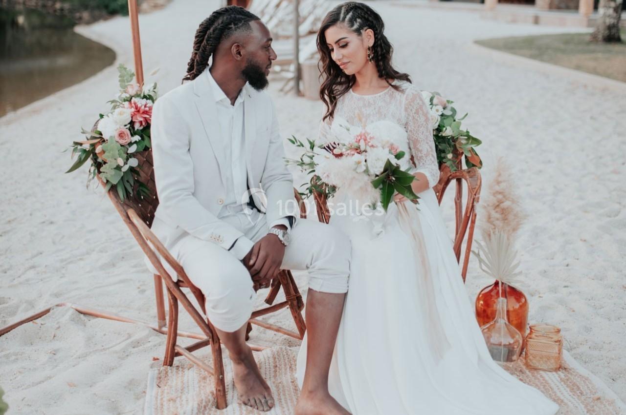 Un couple en tenue de mariage assis sur des chaises décorées de fleurs, sur une plage de sable.