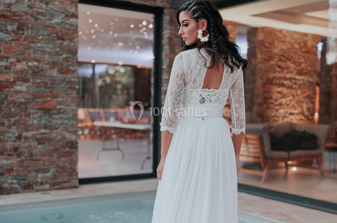 Femme en robe de mariée en dentelle blanche, debout près d'une piscine, avec un intérieur moderne en arrière-plan.