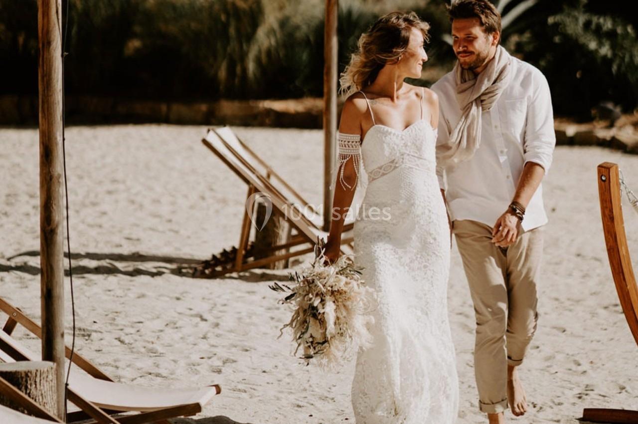 Un couple marche sur une plage ensoleillée, la femme en robe blanche tenant un bouquet, entourés de chaises longues en bois.
