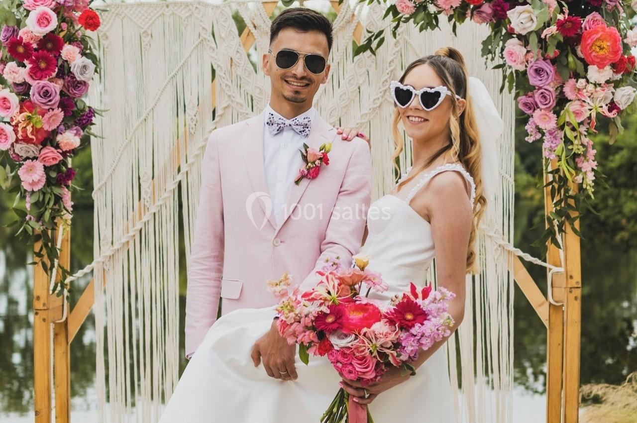 Un couple souriant en tenue de mariage pose devant une arche décorée de fleurs colorées et de macramé.