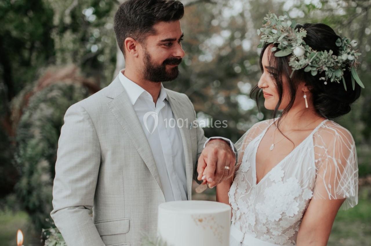 Un couple en tenue de mariage se regarde avec complicité près d'un gâteau, dans un cadre naturel verdoyant.