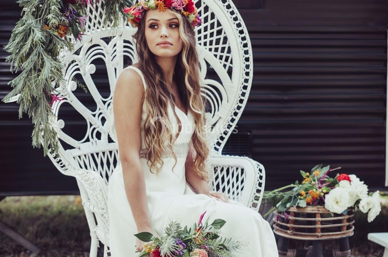 Femme en robe blanche assise sur une chaise en osier décorée de fleurs, tenant un bouquet coloré.
