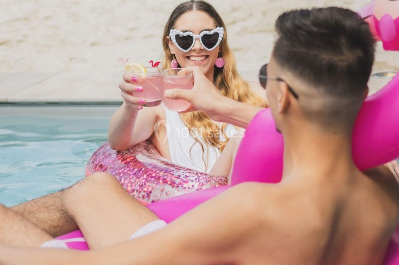 Une femme souriante dans une bouée rose trinque avec un homme dans une piscine, en arrière-plan une plage.