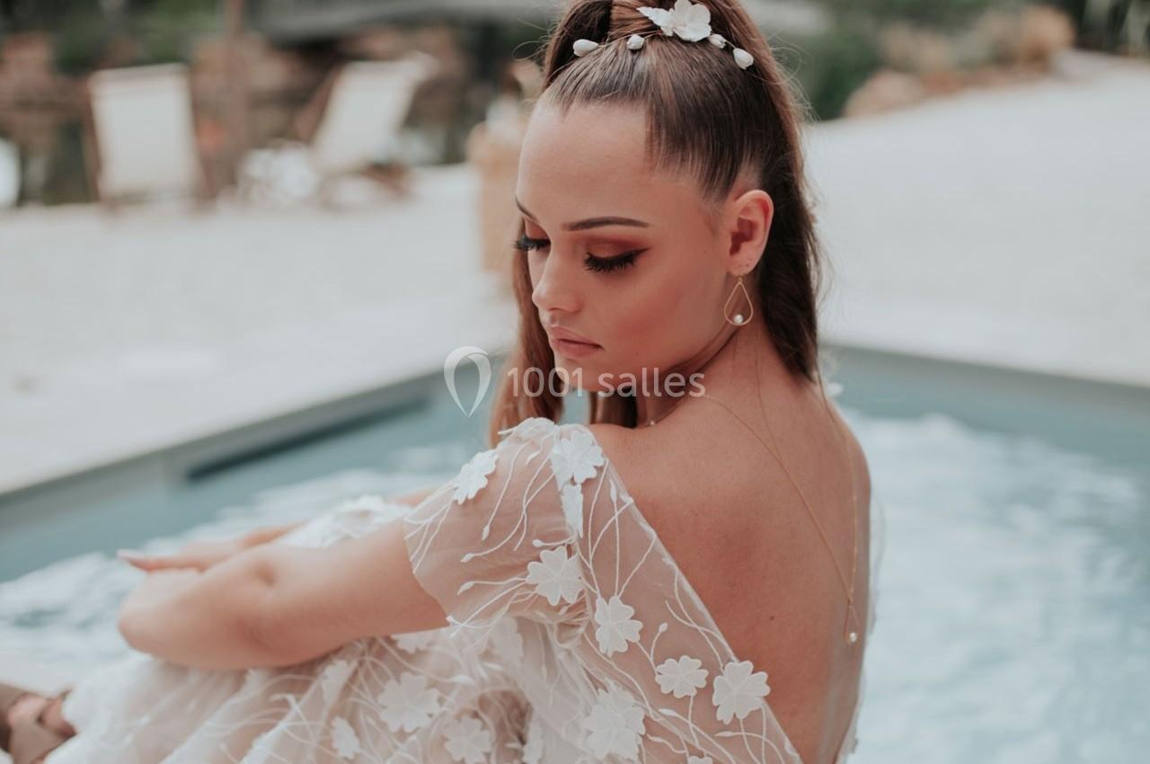 Une femme en robe blanche ornée de fleurs assise près d'une piscine, coiffée d'un chignon décoré de fleurs.