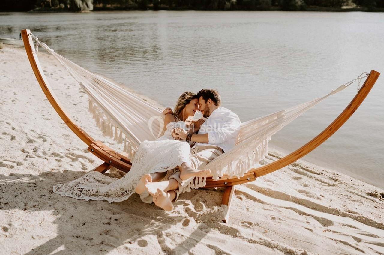 Un couple allongé dans un hamac en bois sur une plage, près d'un lac, partage un moment de tendresse.