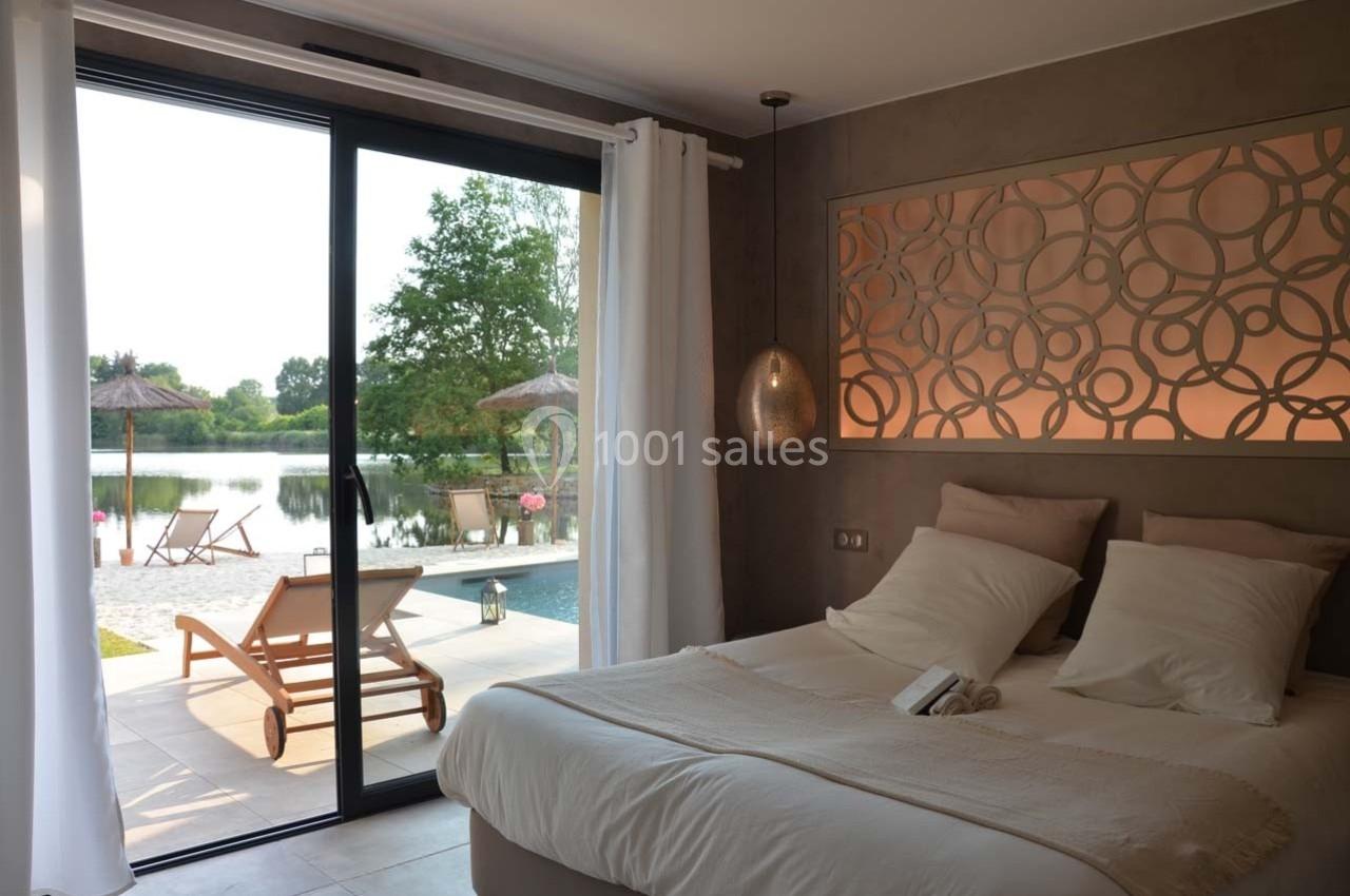 Chambre lumineuse avec lit double, grande baie vitrée donnant sur une terrasse, piscine et vue sur un lac.