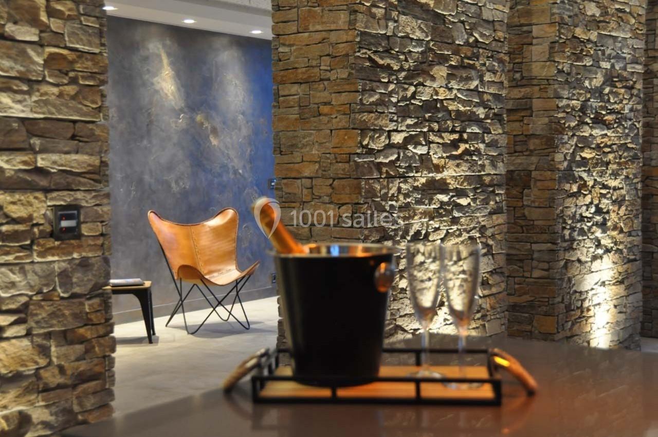 Mur en pierre éclairé, fauteuil en cuir et seau à champagne avec verres sur une table au premier plan.