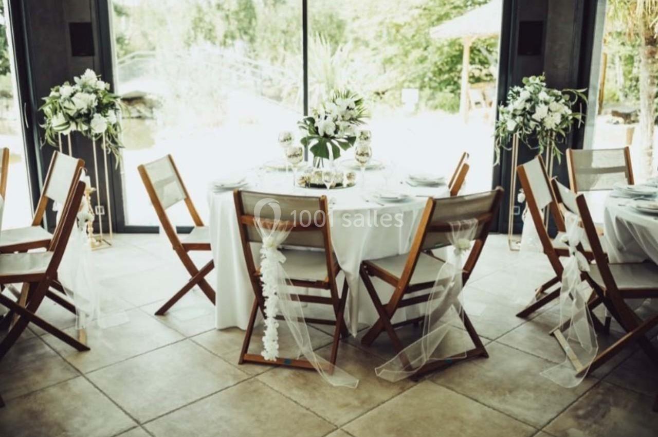 Table ronde décorée pour un événement, entourée de chaises en bois avec voilages, située près de grandes baies vitrées.