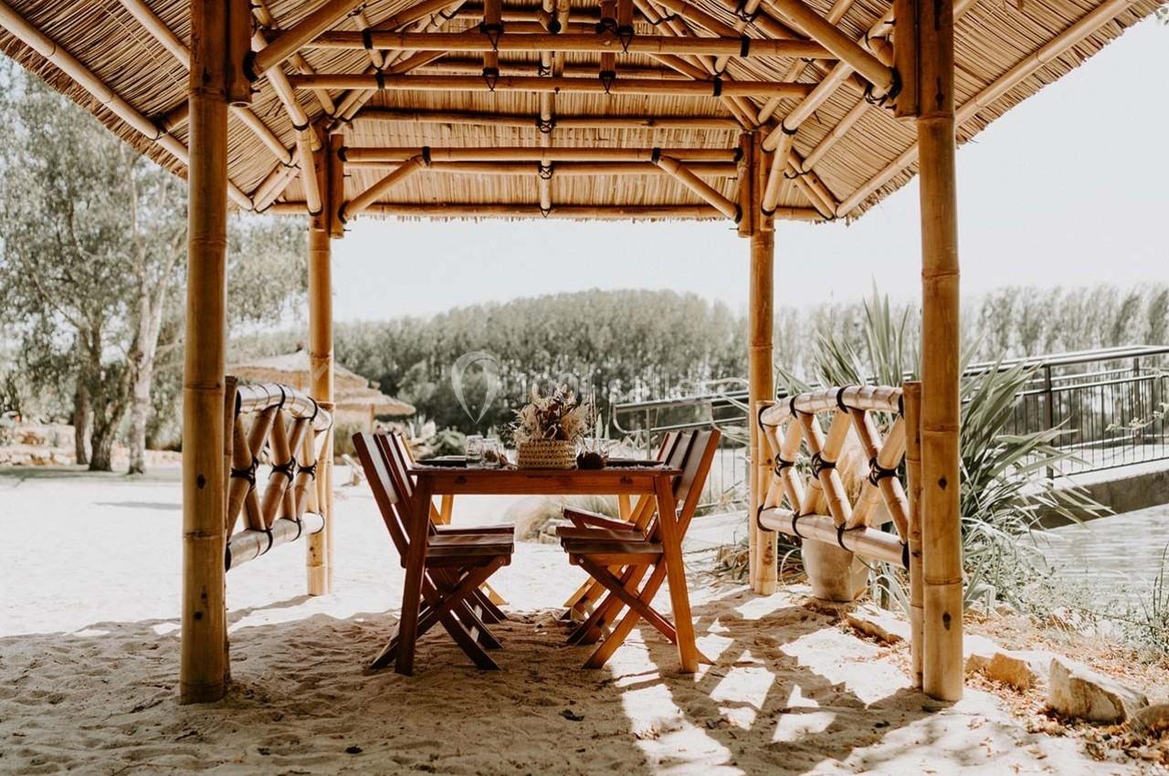 Abri en bambou avec table et chaises en bois sur un sol sablonneux, entouré de végétation et proche d'un pont.