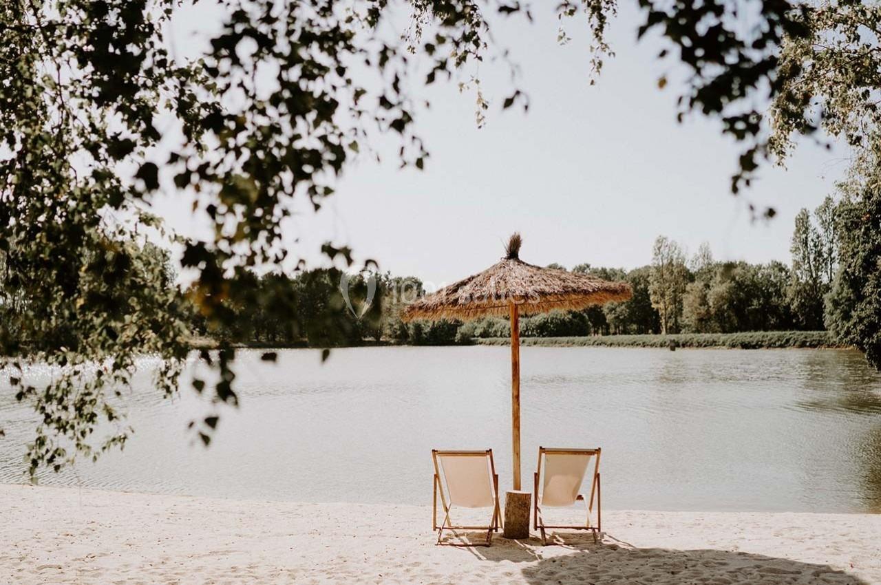 Deux chaises longues sous un parasol en paille sur une plage au bord d'un lac entouré de végétation.
