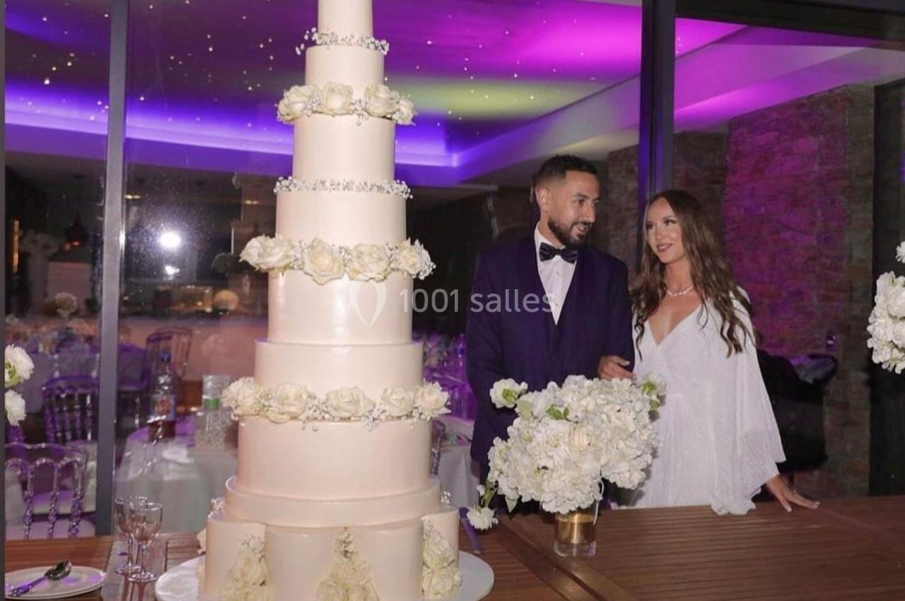 Un couple en tenue de soirée pose près d'un gâteau de mariage à plusieurs étages décoré de fleurs blanches.