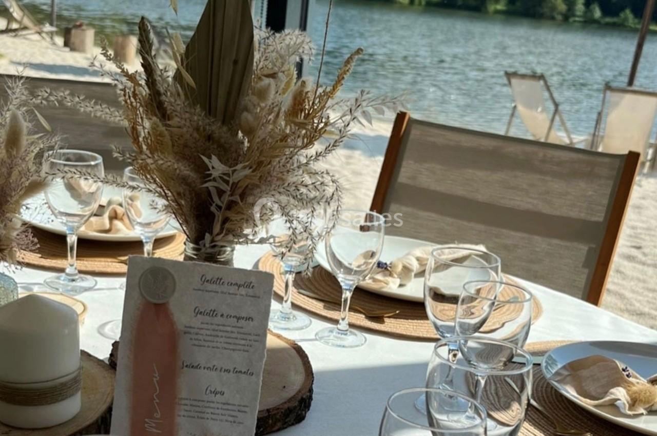 Table dressée en extérieur avec décoration naturelle, vue sur un lac, chaises longues en arrière-plan.