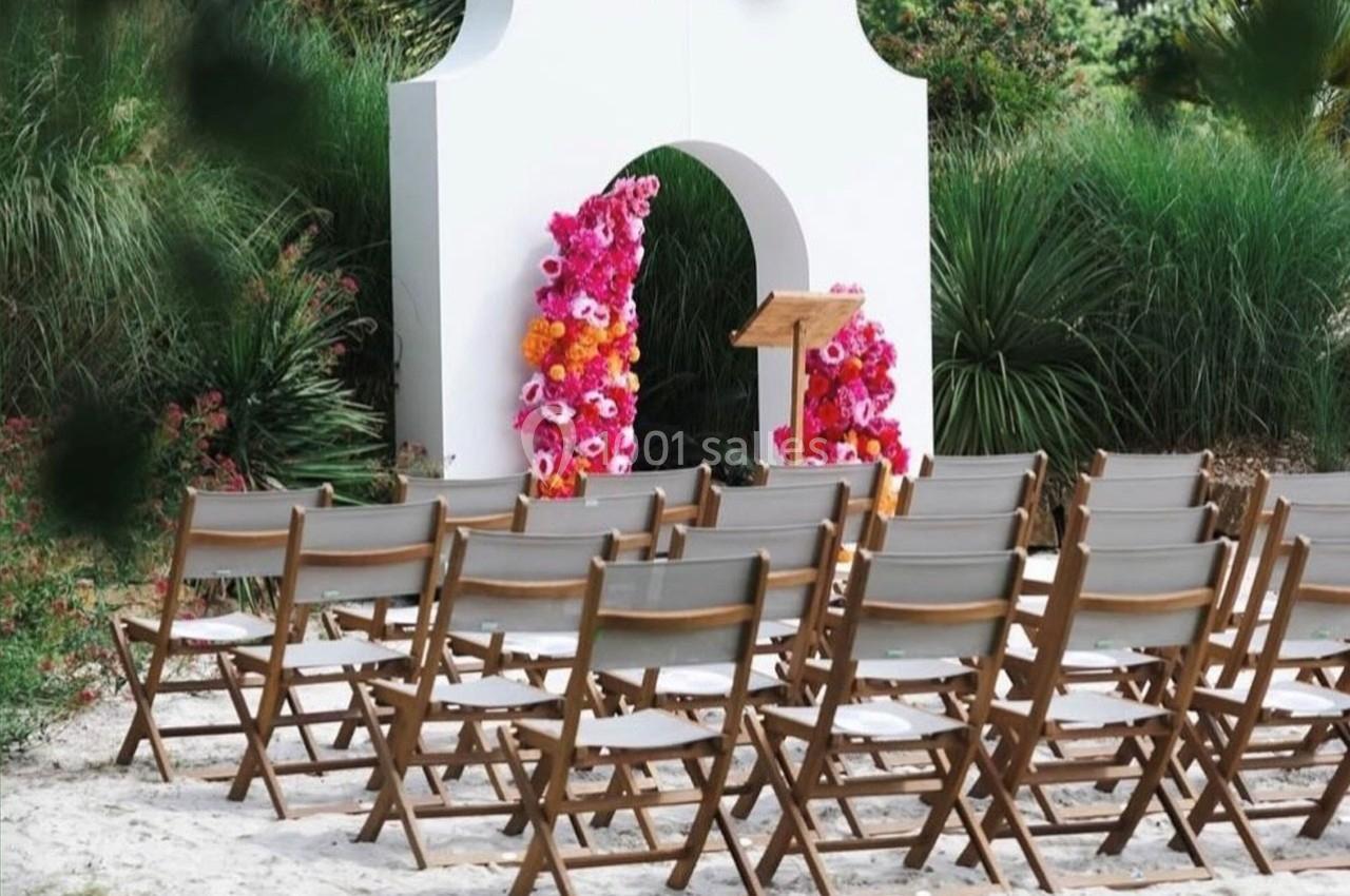 Chaises en bois disposées devant une arche blanche décorée de fleurs colorées dans un cadre extérieur verdoyant.