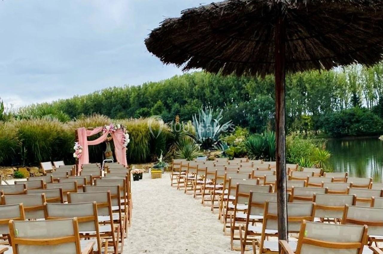 Chaises en bois disposées face à une arche décorée de fleurs, sur une plage bordée de végétation et d'un plan d'eau.