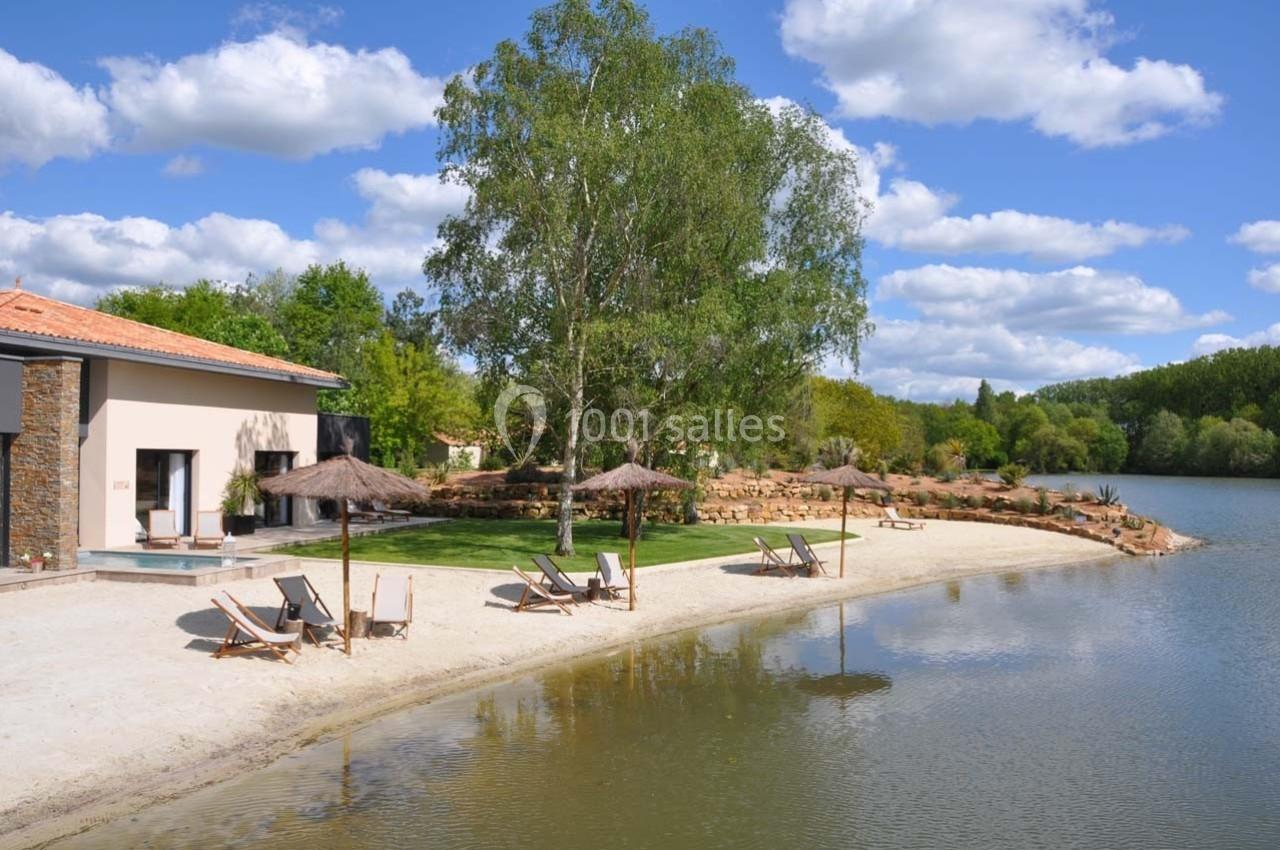 Plage aménagée avec transats et parasols au bord d’un étang, entourée de verdure et d’un bâtiment moderne.