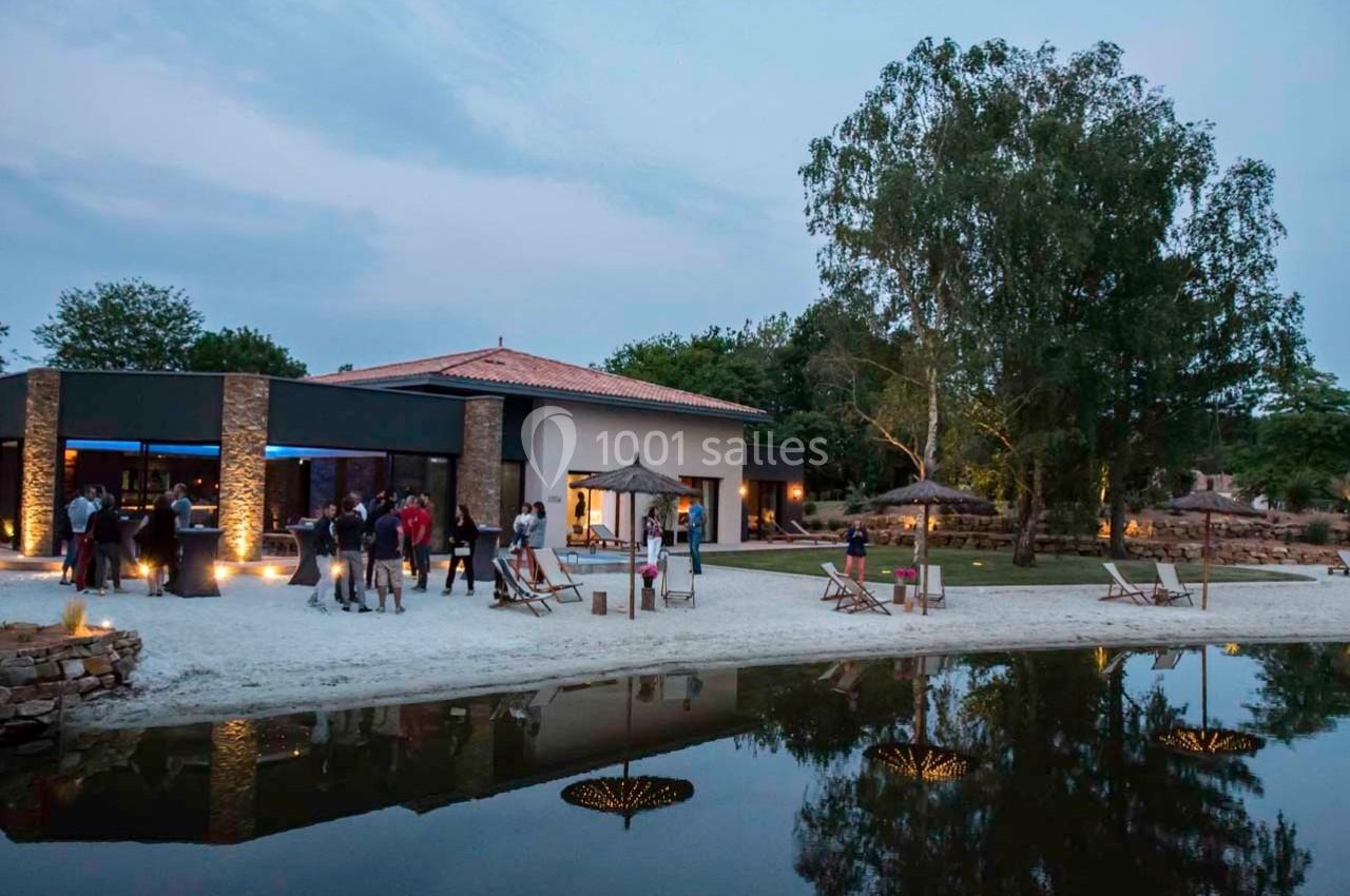 Vue d'un bâtiment moderne entouré de verdure, avec des personnes rassemblées près d'un étang au crépuscule.