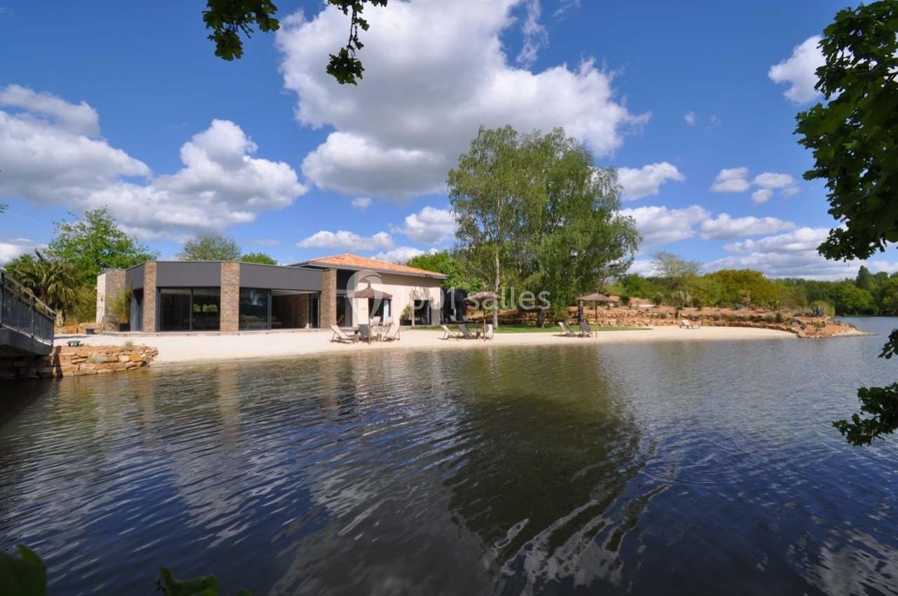 Maison moderne avec terrasse au bord d'un étang, entourée de végétation sous un ciel bleu parsemé de nuages.