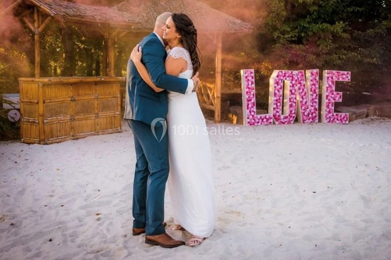 Un couple s'enlace sur une plage, devant une décoration en bambou et le mot ’LOVE’ formé de fleurs roses.