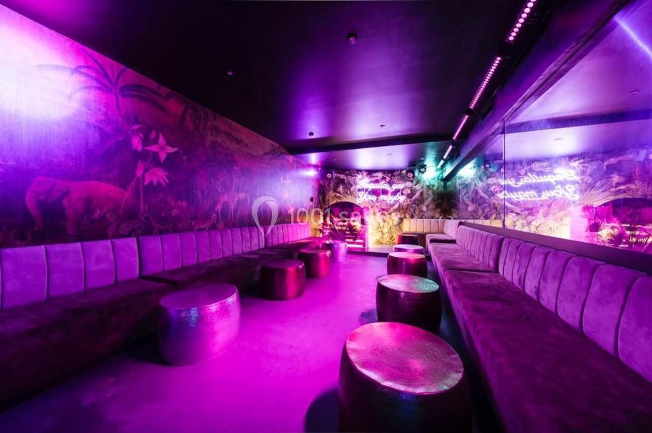 Salle lounge avec banquettes violettes, tables basses métalliques et éclairage néon rose dans une ambiance tropicale.