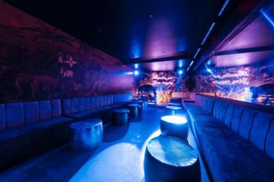 Salle sombre avec éclairage tamisé rouge, mobilier élégant, chandeliers et étagères illuminées derrière un bar.