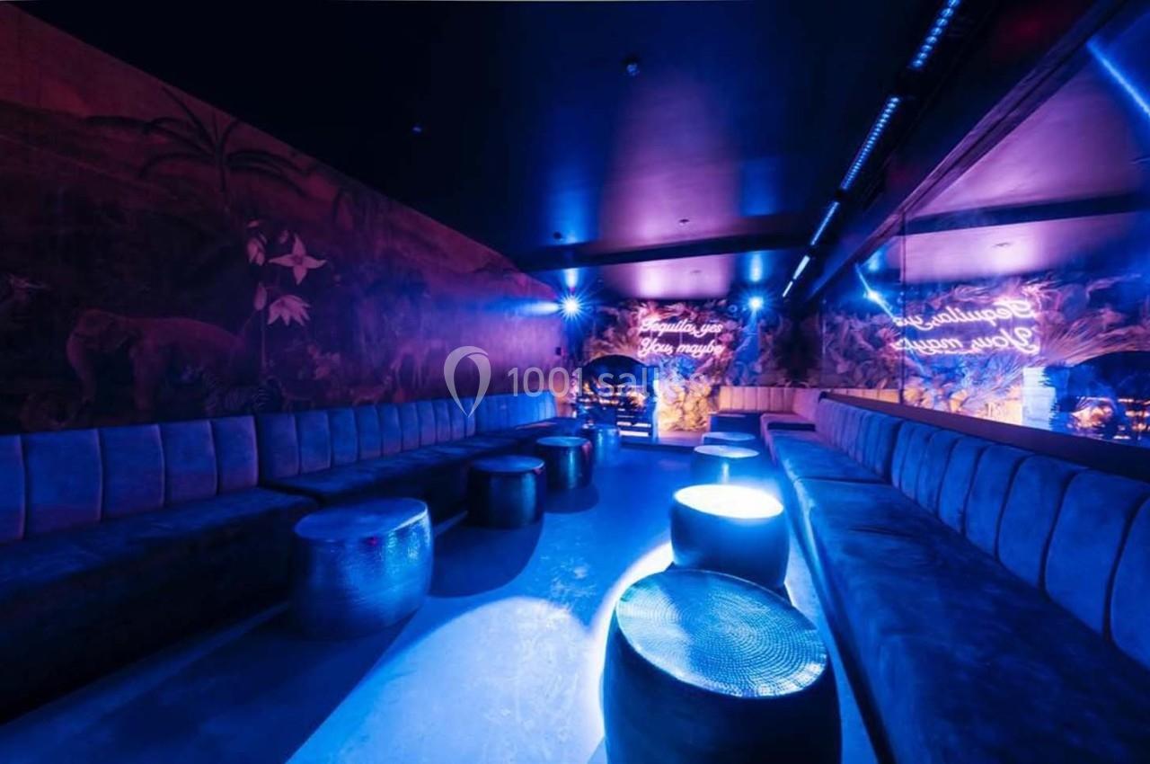 Salle lounge avec banquettes et tables basses, éclairée par des lumières bleues et des néons décoratifs au mur.