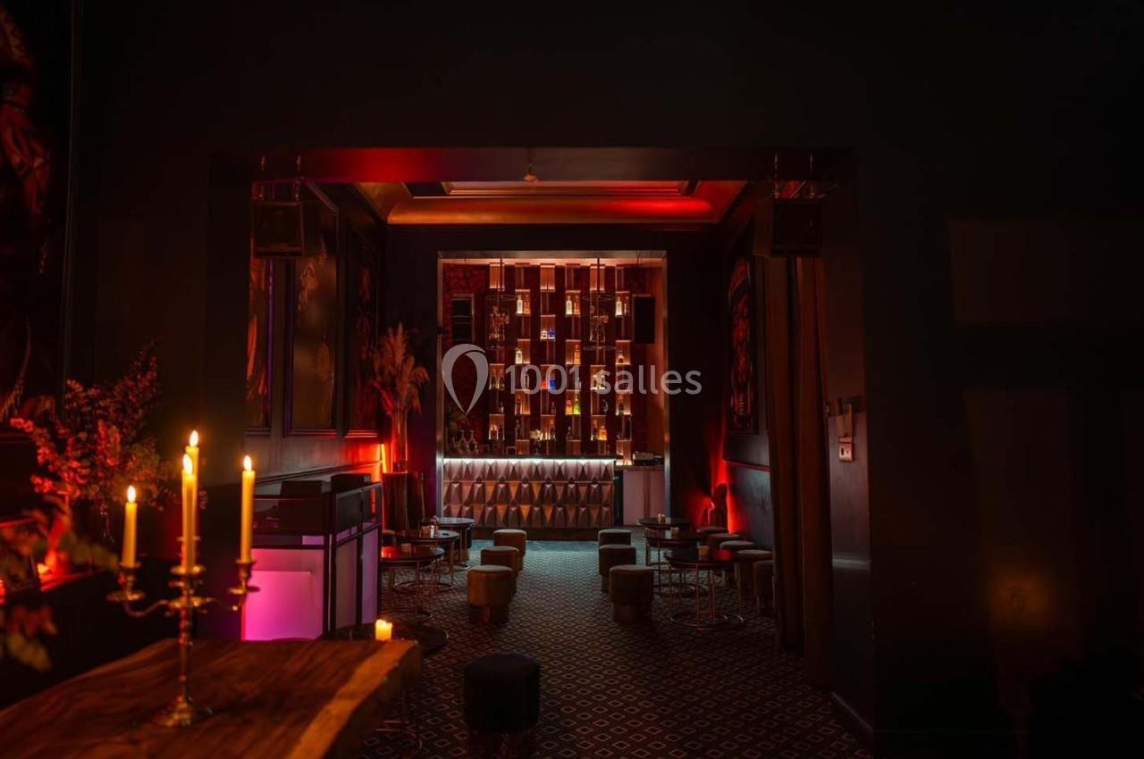 Salle sombre et élégante avec éclairage tamisé, bar illuminé en arrière-plan et chandeliers sur une table au premier plan.