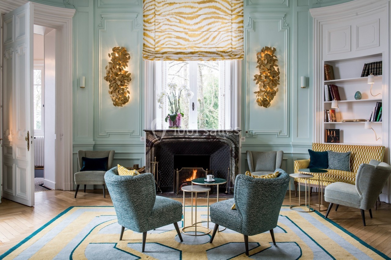 Salon élégant avec cheminée allumée, fauteuils bleus, table basse ronde et décoration murale dorée.