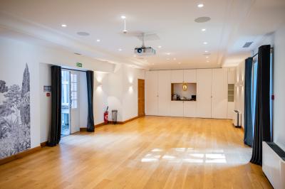 Salle lumineuse avec parquet en bois, murs blancs, rideaux sombres, projecteur au plafond et portes vitrées donnant sur l…