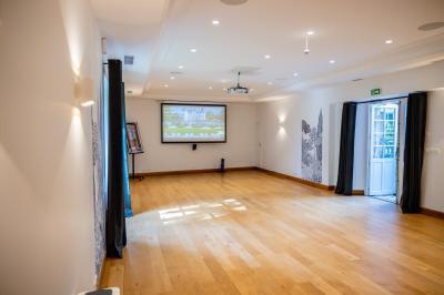 Salle lumineuse avec parquet en bois, murs blancs, rideaux sombres, projecteur au plafond et portes vitrées donnant sur l…