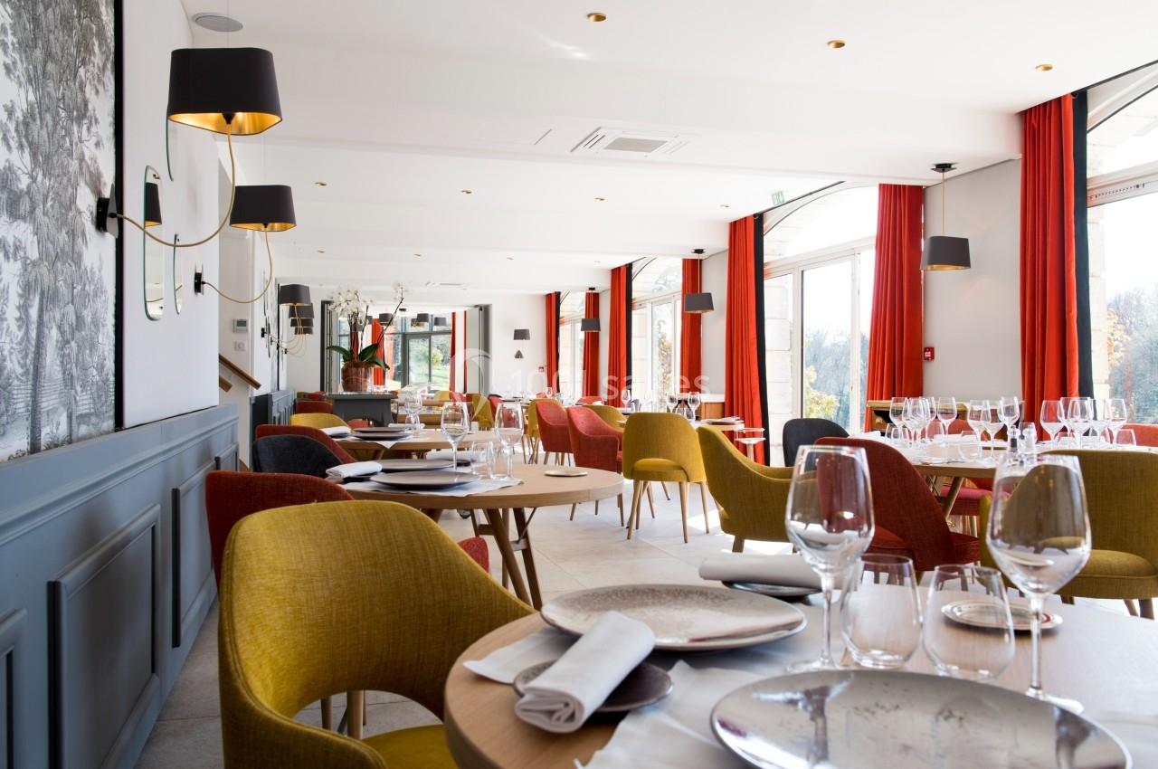 Salle de restaurant lumineuse avec tables dressées, chaises colorées et grandes baies vitrées laissant entrer la lumière…