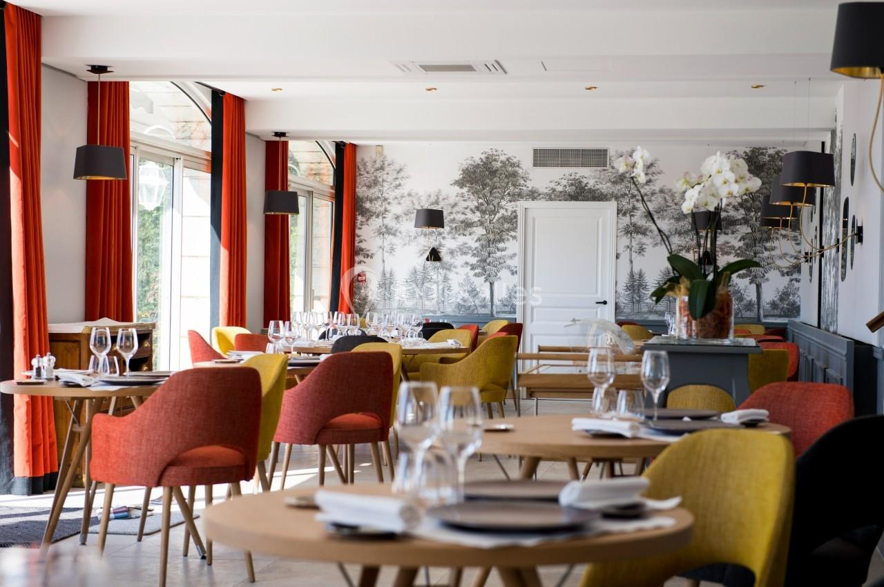 Salle de restaurant lumineuse avec tables dressées, chaises colorées et décoration murale représentant un paysage forestier.