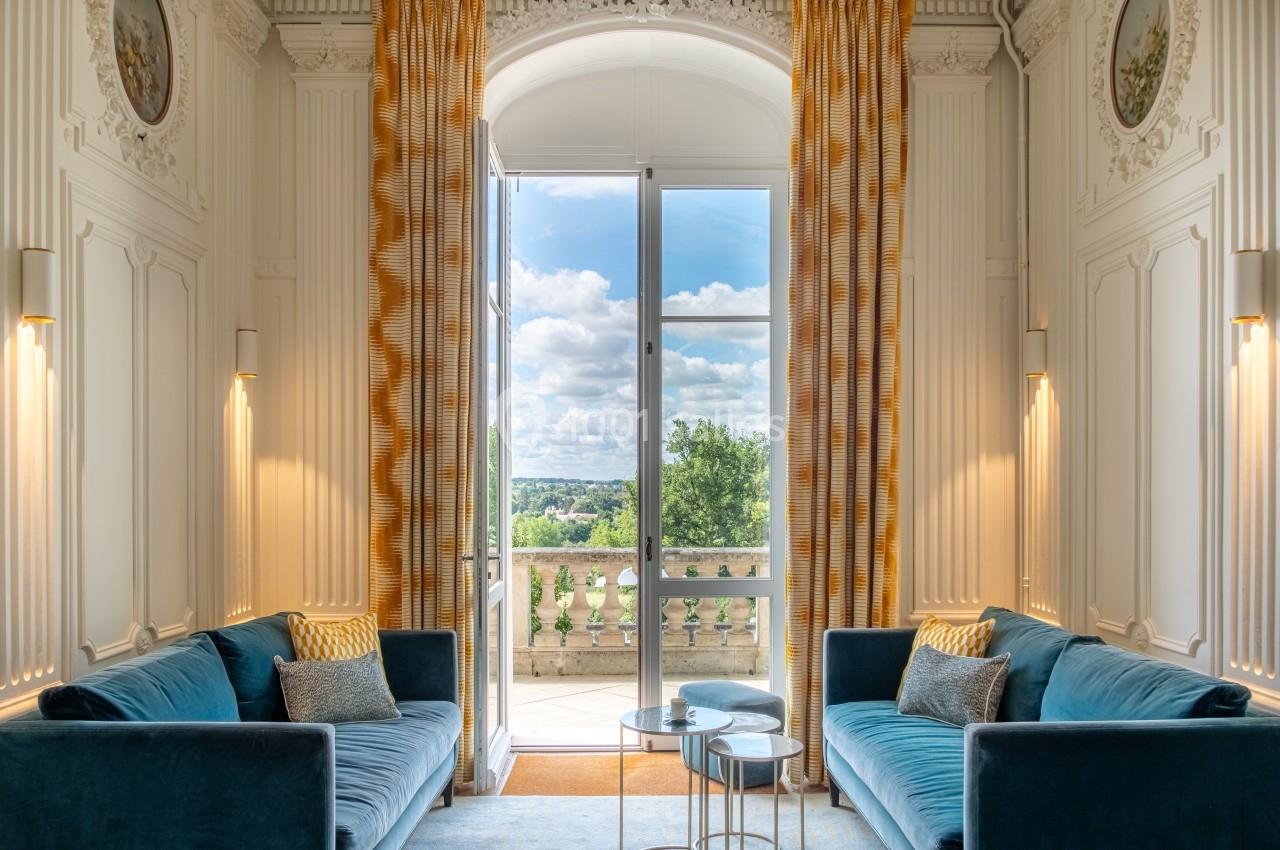Salon lumineux avec deux canapés bleus, tables basses et grandes fenêtres ouvertes sur un balcon avec vue sur la campagne.