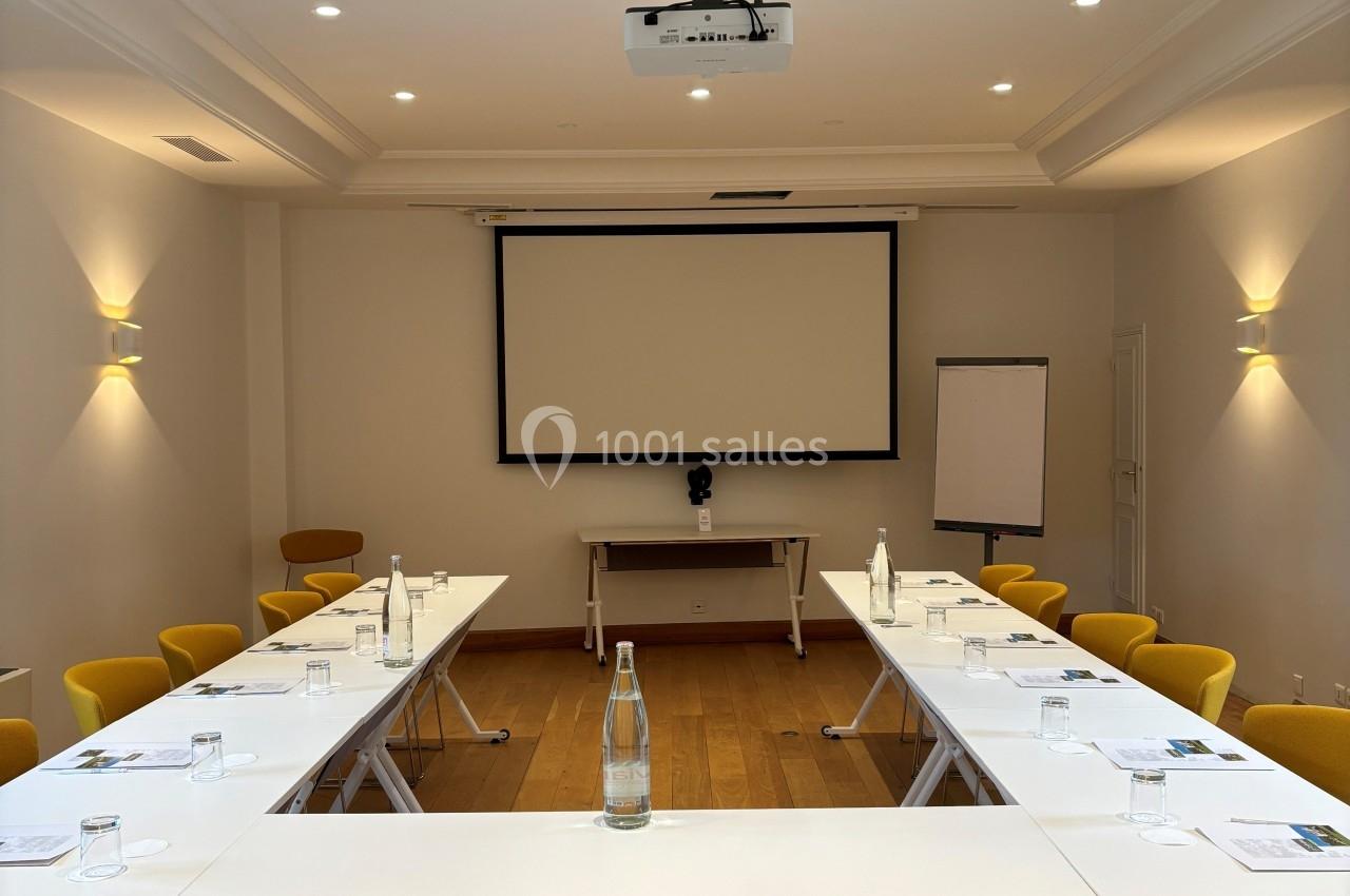 Salle de réunion lumineuse avec tables disposées en U, chaises jaunes, écran de projection et tableau blanc.