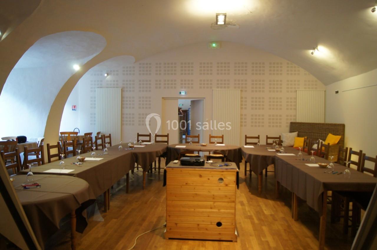 Salle de réunion avec tables disposées en U, chaises en bois, projecteur sur un meuble et éclairage au plafond.