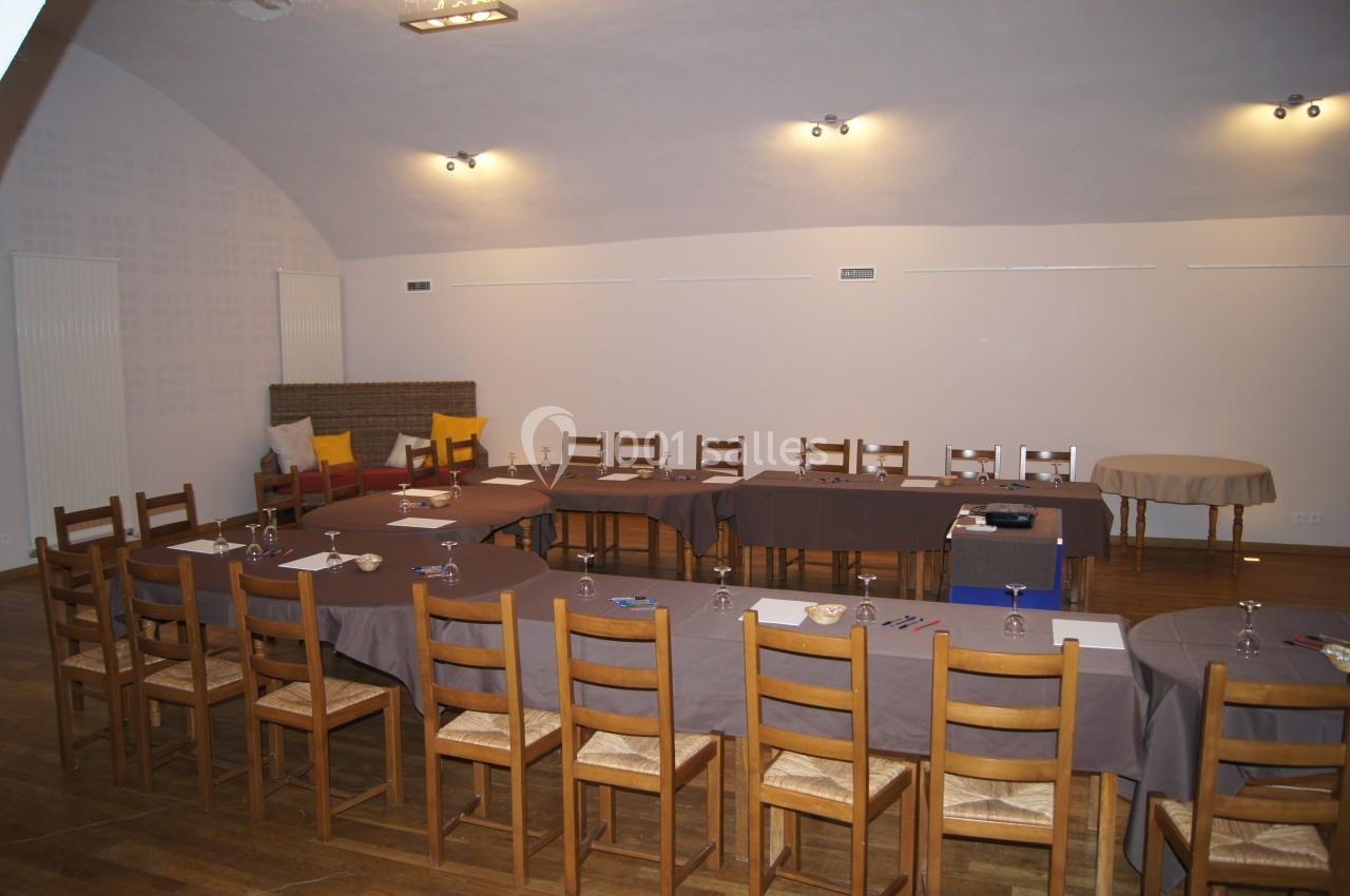 Salle de réunion aménagée en U avec des tables recouvertes de nappes grises, chaises en bois et matériel de travail.